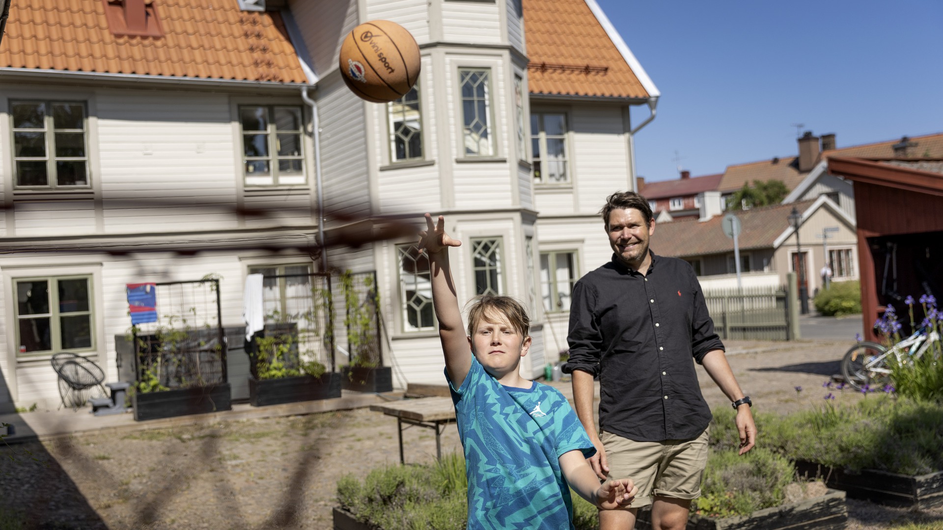 Far och son spelar basket i trädgården, med en stor trävilla i bakgrunden.